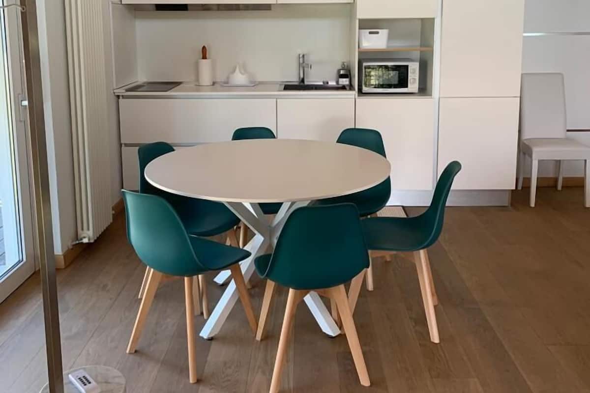 A modern kitchen with white cabinets, a round table, teal chairs, and a hanging light fixture.