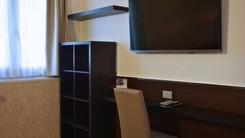 Une chambre d’hôtel dotée d’un grand téléviseur à écran plat fixé au mur, d’un meuble de couleur sombre, d’un fauteuil beige et d’une petite table sur laquelle se trouve une télécommande.