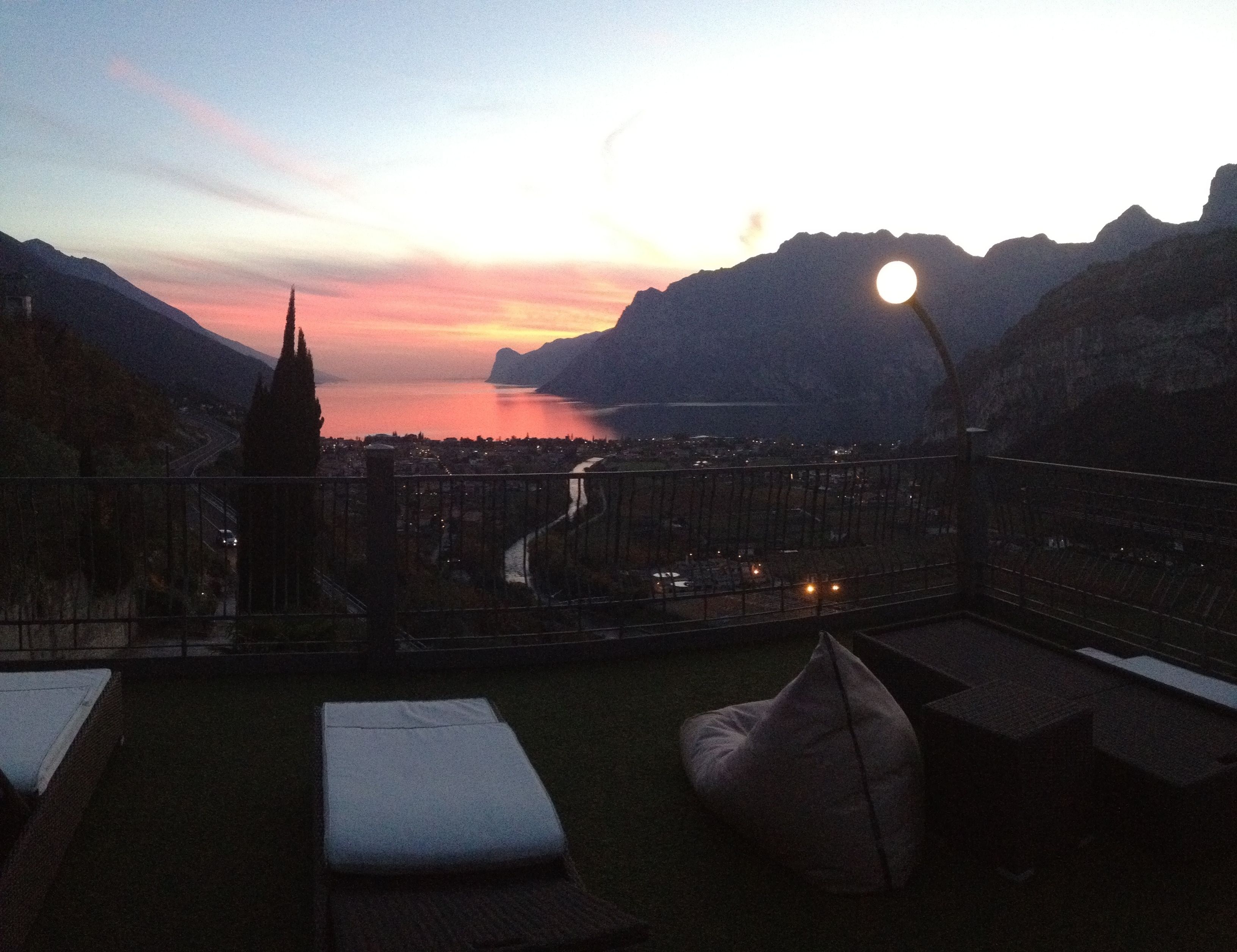 Un balcon offrant une vue sur un coucher de soleil se reflétant sur une étendue d’eau, le tout étant entouré de montagnes.