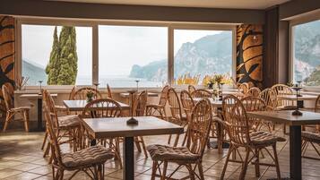 Un restaurant avec des chaises et des tables en osier, de grandes fenêtres offrant une vue sur la mer et les montagnes.