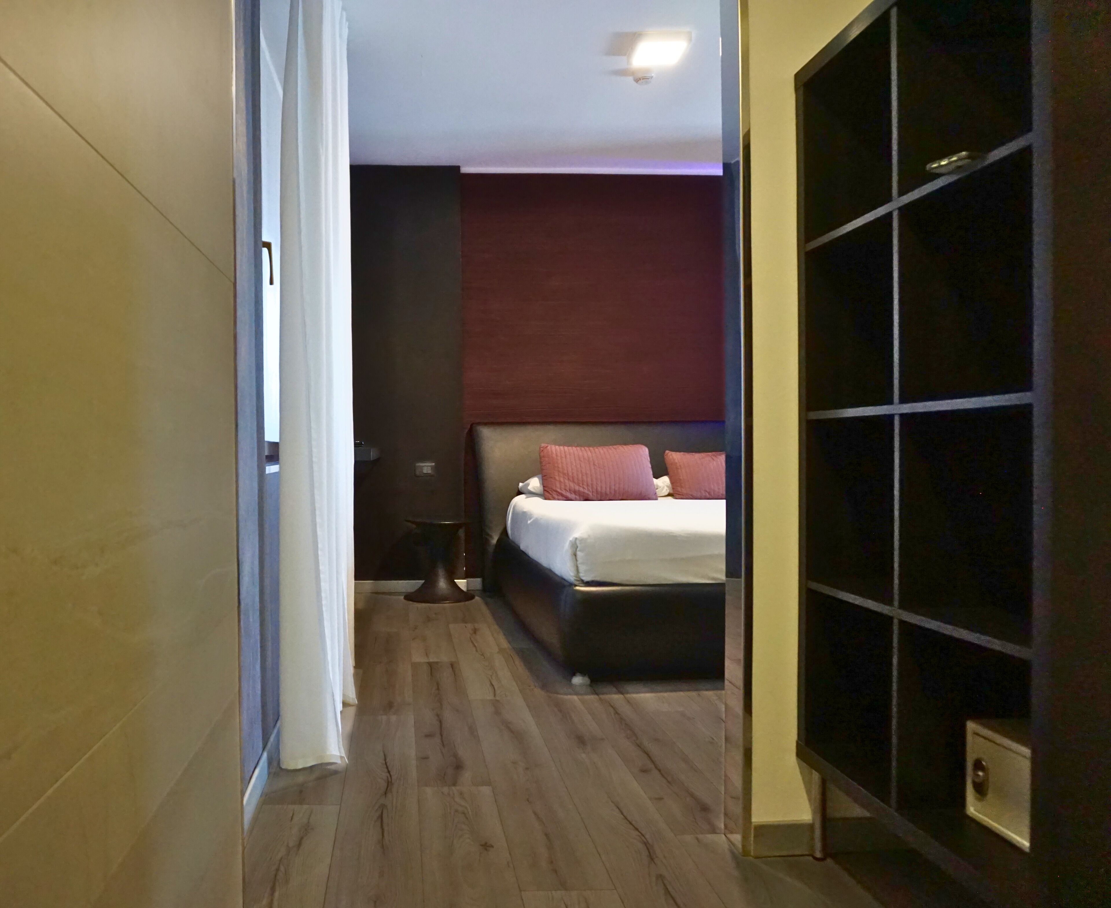 Ein Hotelzimmer mit Holzboden, einem Bett mit roten Kissen, einem Nachttisch und einem großen, schwarzen Kleiderschrank.