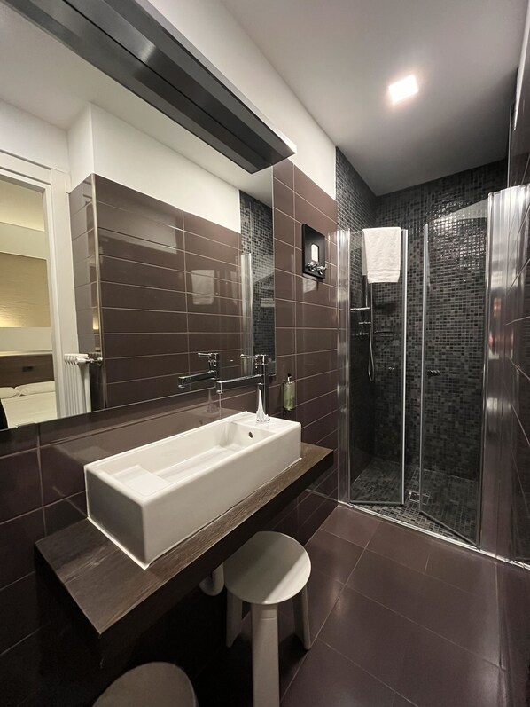 Un baño moderno con un lavamanos grande de color blanco, un espejo, cabina de ducha de cristal y paredes cubiertas de baldosas marrón oscuro.