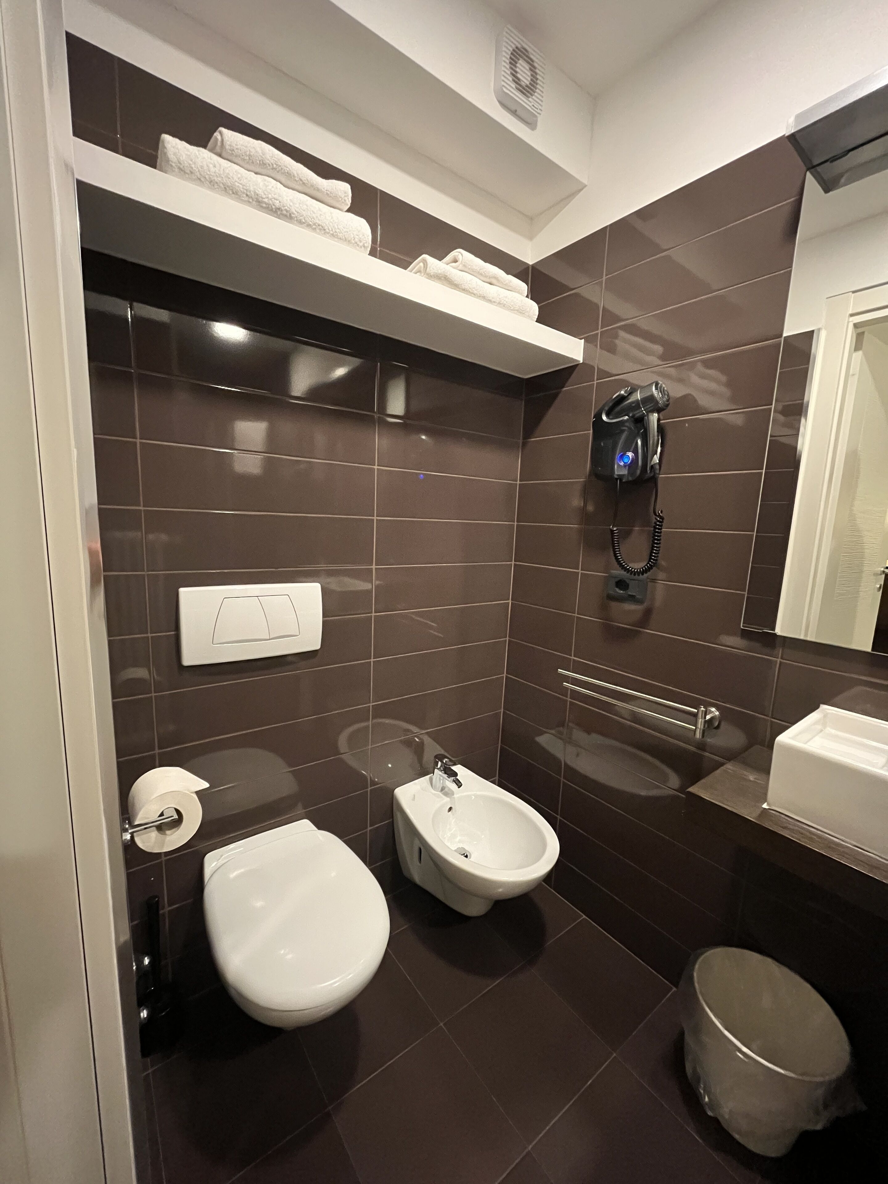 Un bagno moderno con piastrelle marrone scuro, un water, un bidet, un lavandino, un asciugacapelli fissato alla parete e un cestino.