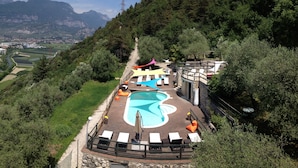Zona de piscina amb gandules i ombrel·les, envoltada d'arbres i un paisatge de muntanyes.