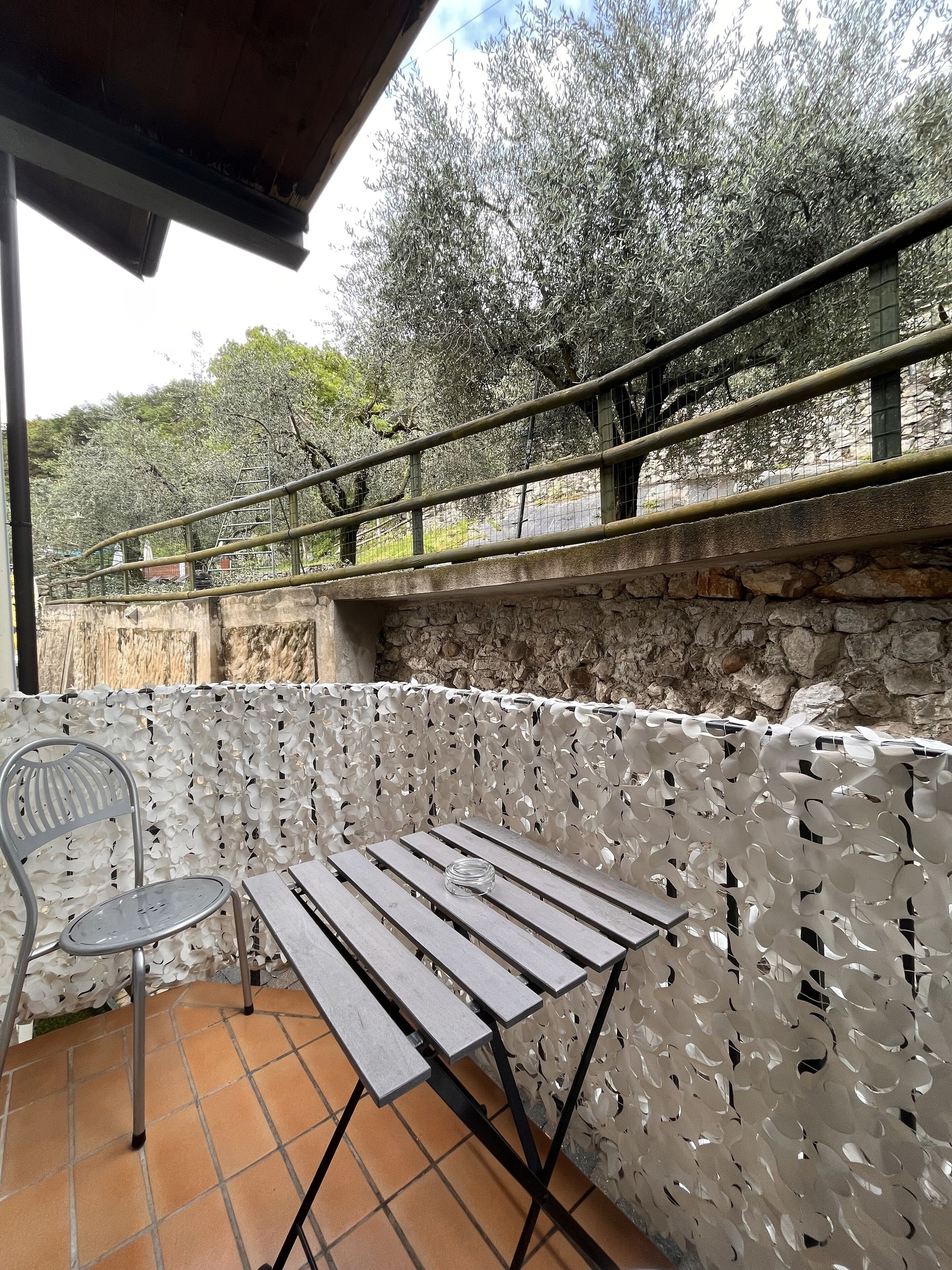 An outdoor patio with a wooden table and a metal chair, a stone wall, and a metal railing overlooking a garden with trees.