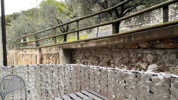 An outdoor patio with a wooden table and a metal chair, a stone wall, and a metal railing overlooking a garden with trees.