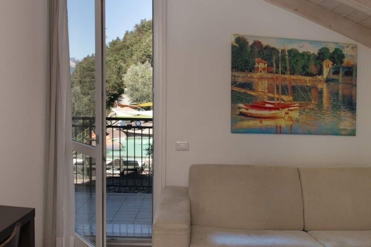 A living room with a beige sofa, a painting of a harbor scene, and a view of a courtyard with a gate and trees.