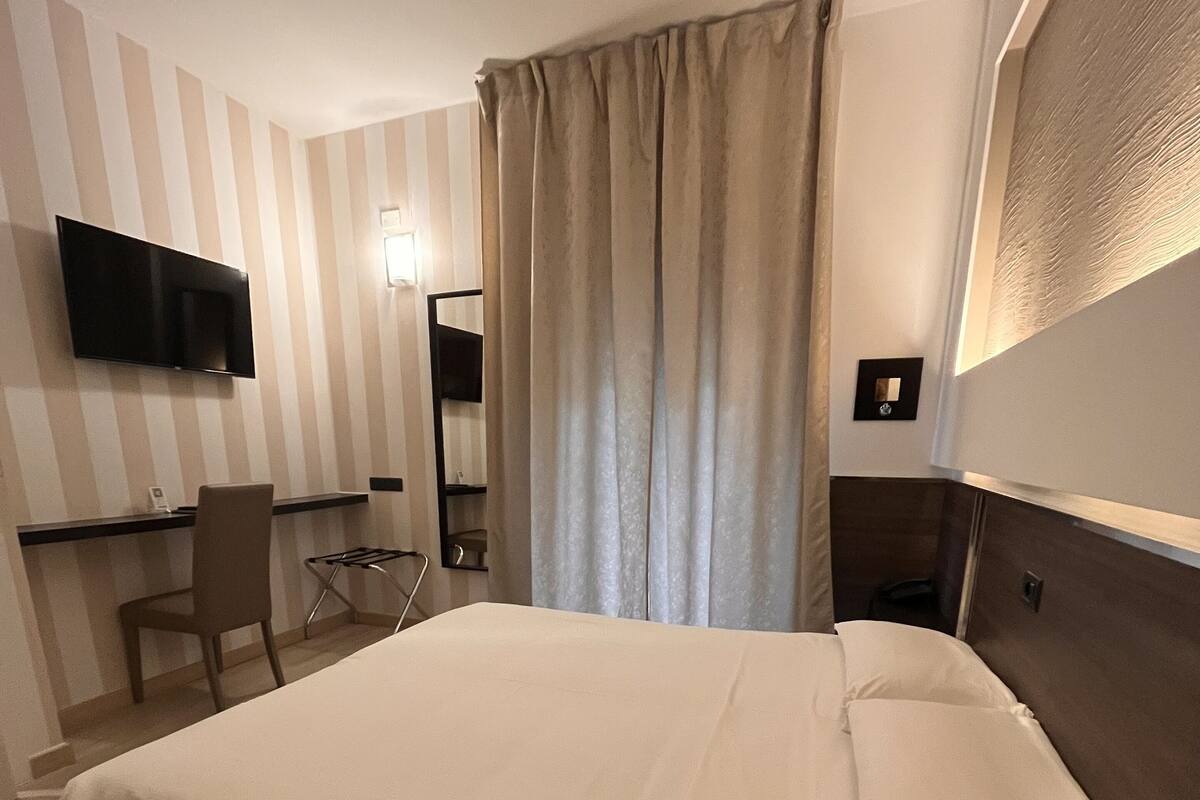 A hotel room with a large bed, a desk with a chair, a TV mounted on the wall, and a window with curtains.