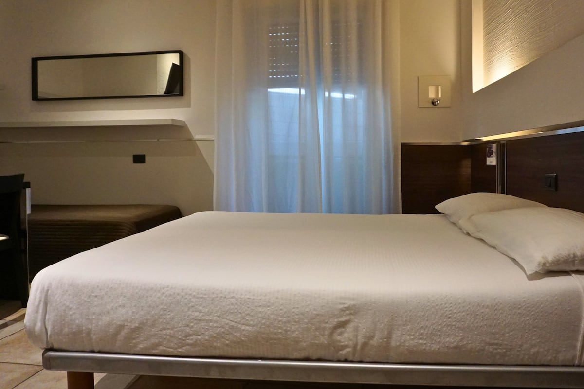 A hotel room with a large bed, a bedside table, a mirror, and a window with sheer curtains.