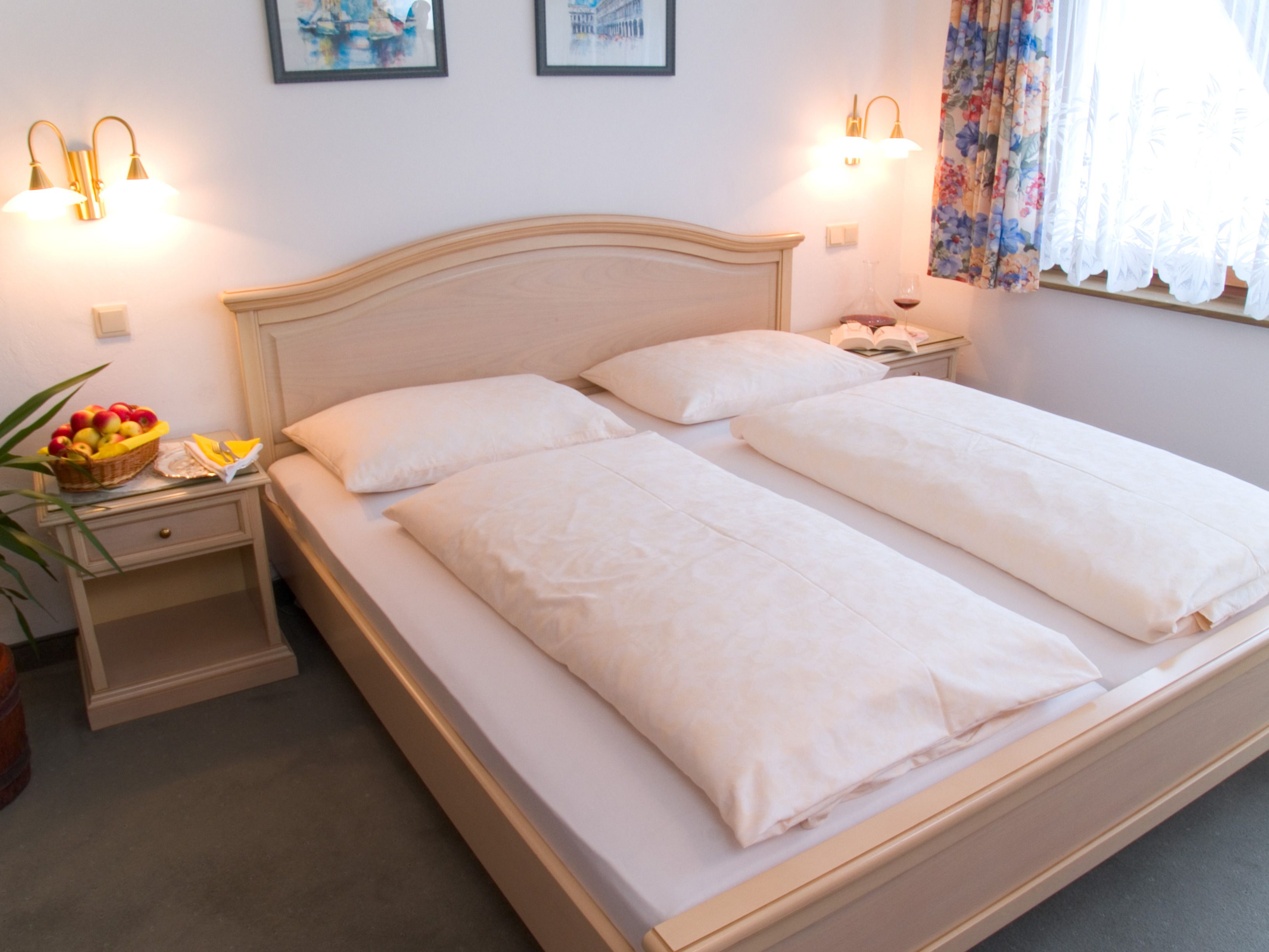 Een netjes opgemaakt bed met witte lakens, een nachtkastje met een fruitmand, en wandlampen.