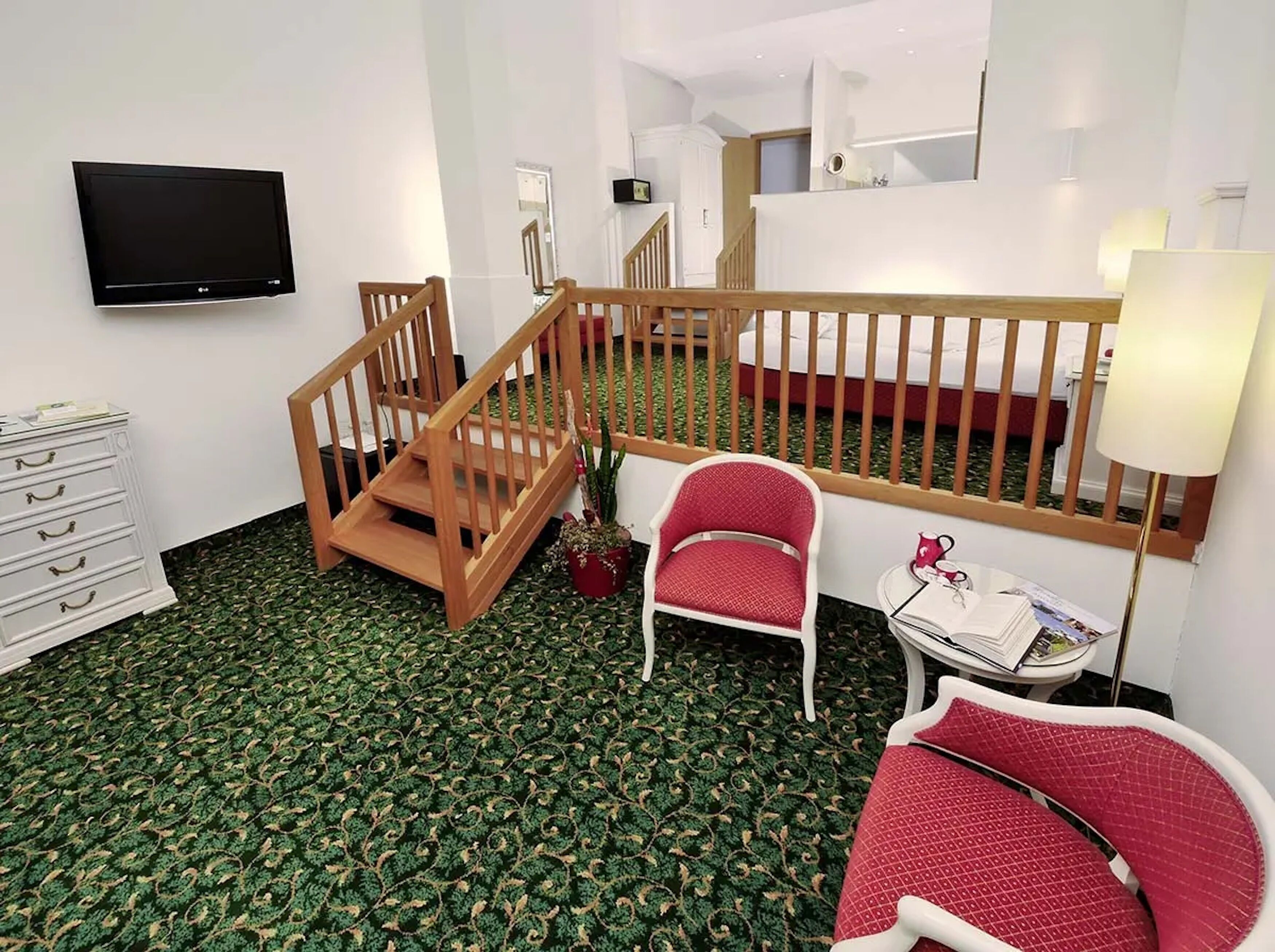 A hotel room with a wooden staircase, a flat-screen TV, a small table with a lamp, and two red chairs.