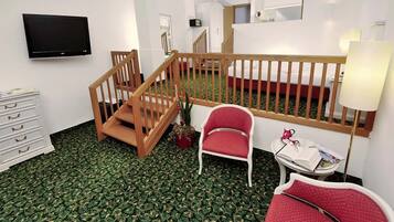 A hotel room with a wooden staircase, a flat-screen TV, a small table with a lamp, and two red chairs.