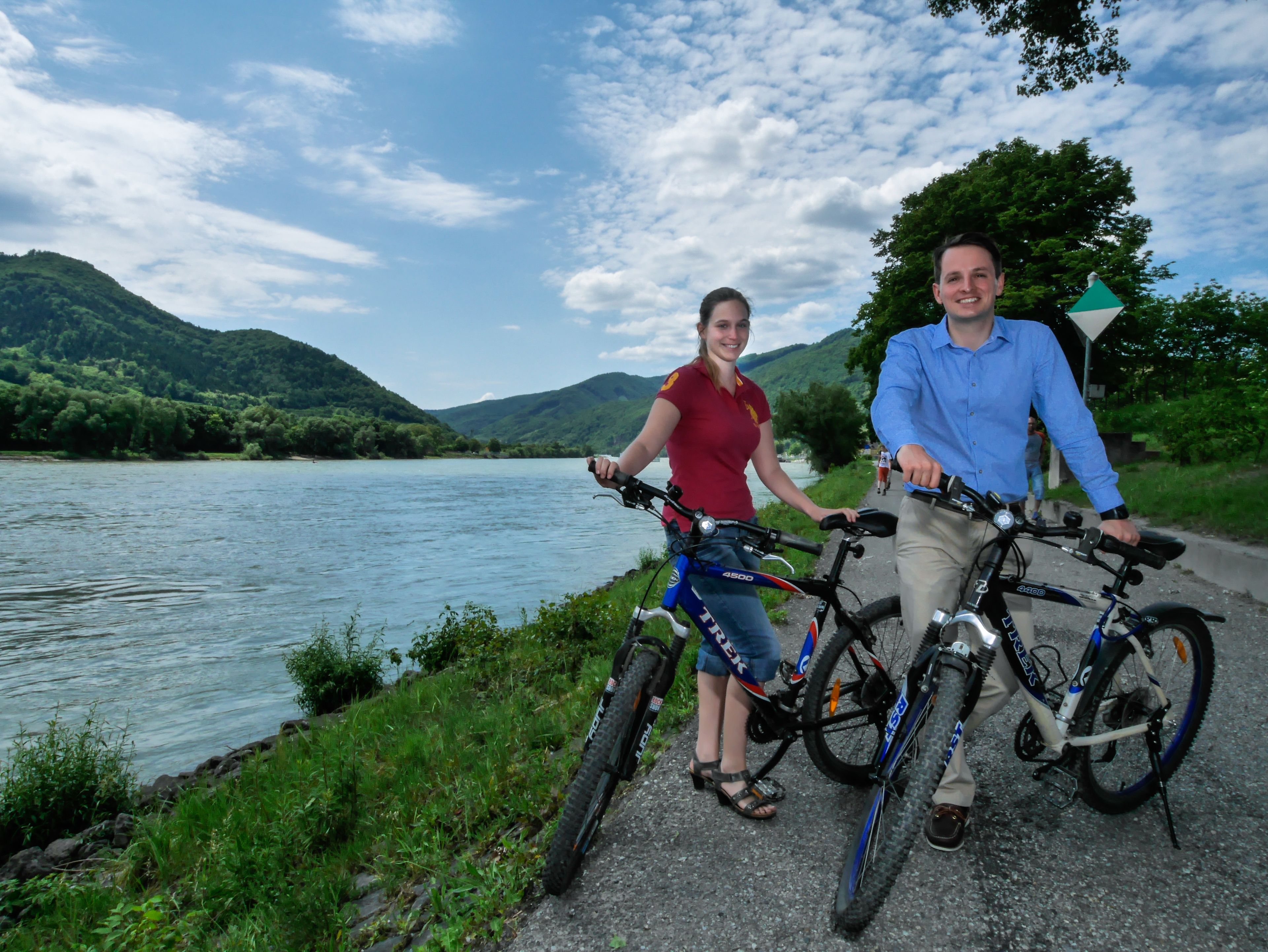 Two people on bicycles by a river with mountains in the background.