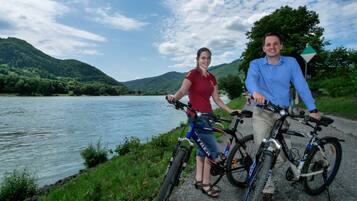Two people on bicycles by a river with mountains in the background.