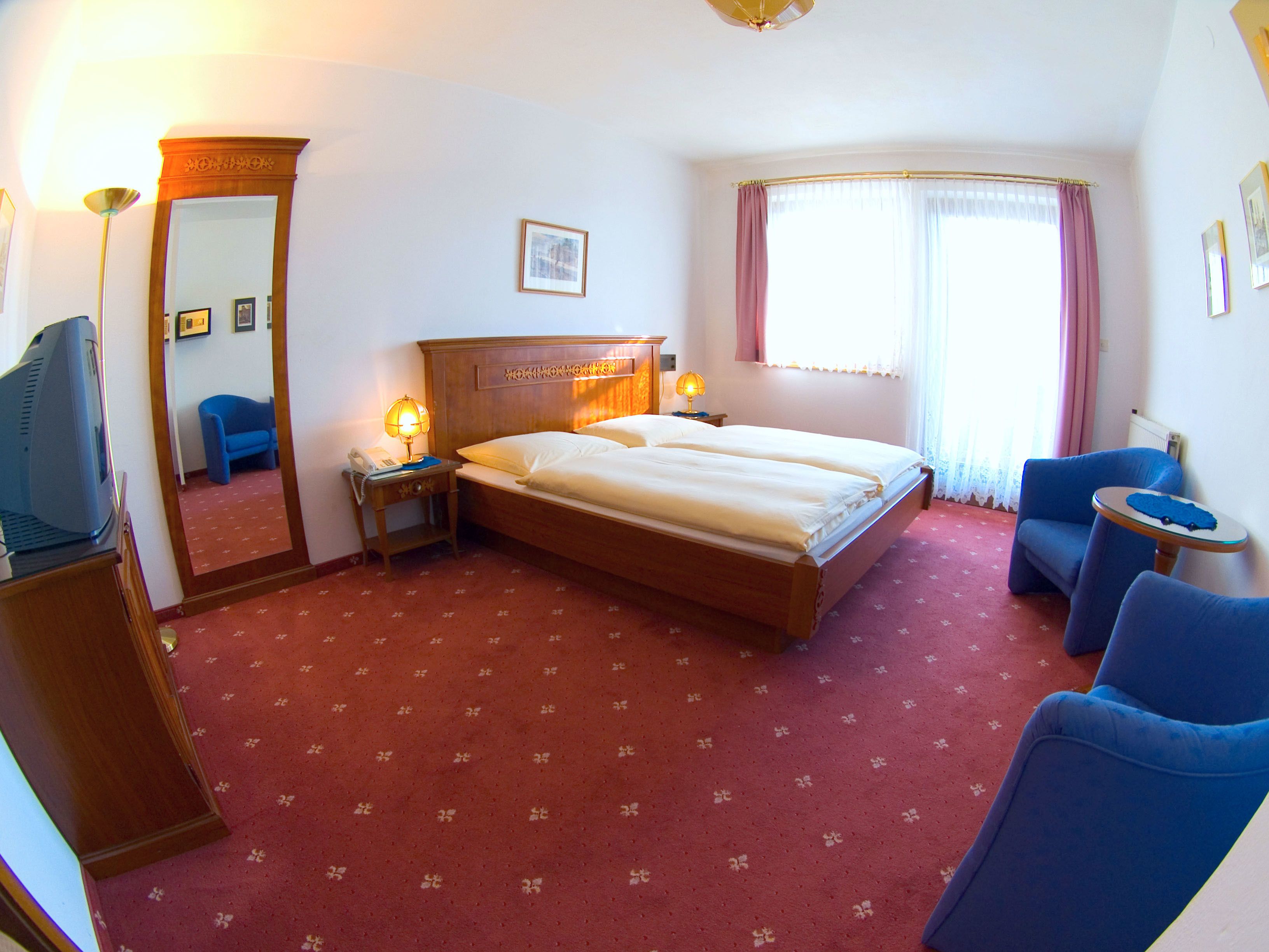 A hotel room with a bed, two bedside tables, a desk with a computer, a chair, a window with curtains, and a mirror.