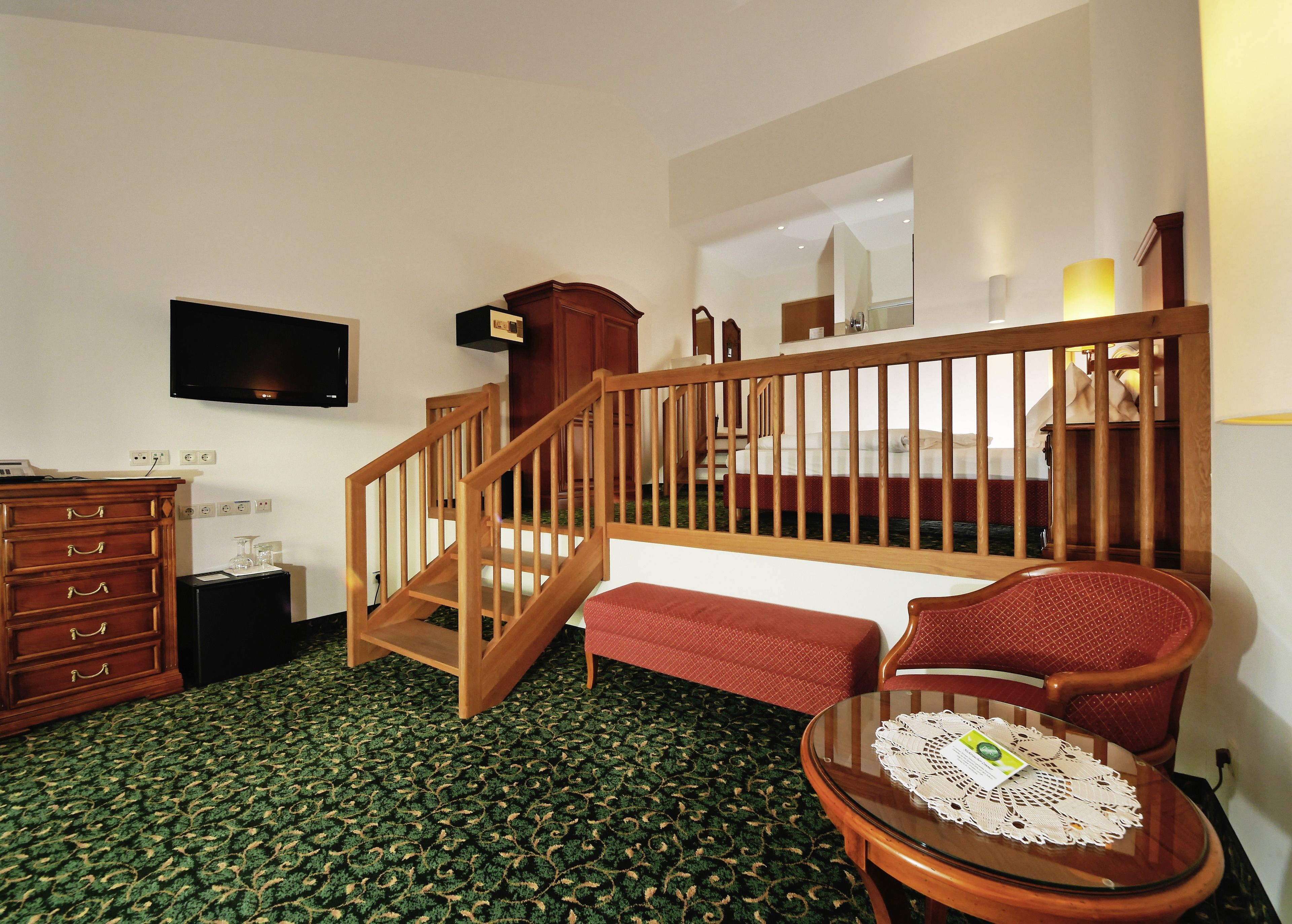 Ein Hotelzimmer mit einer Holztreppe, einem an der Wand befestigten Flachbildfernseher, einer Holzkommode, einem rot gepolsterten Sessel und einem Glastisch mit einer Spitzeneinlage.