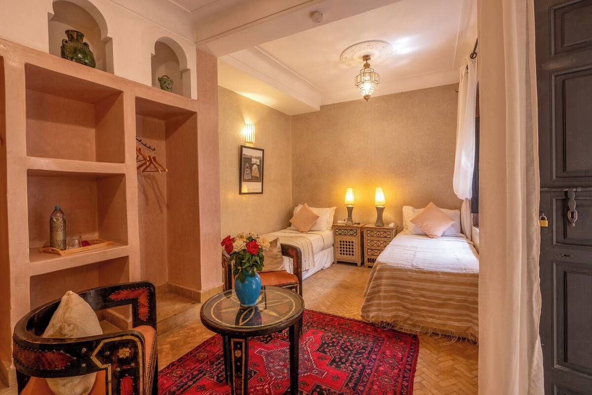 A hotel room with two beds, a sitting area with a chair and table, a red patterned rug, and a decorative vase with flowers.