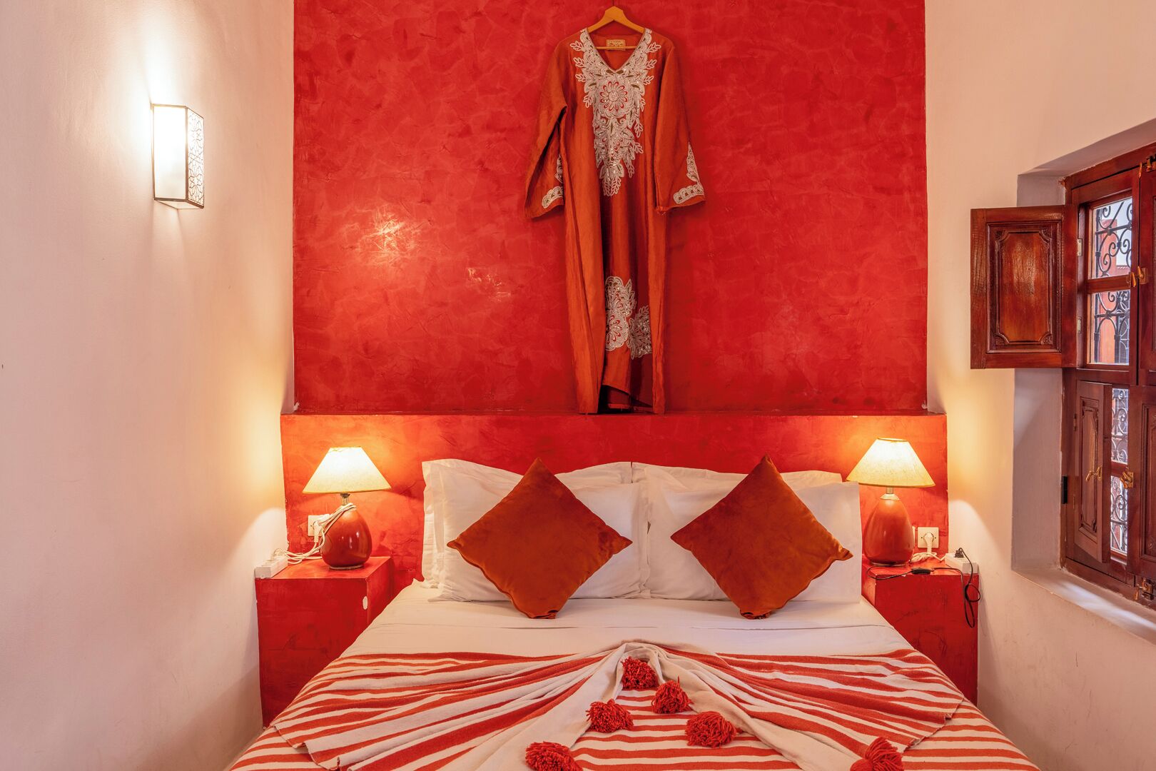 A room with a red accent wall, a bed with striped bedding, two nightstands with lamps, and a hanging robe.