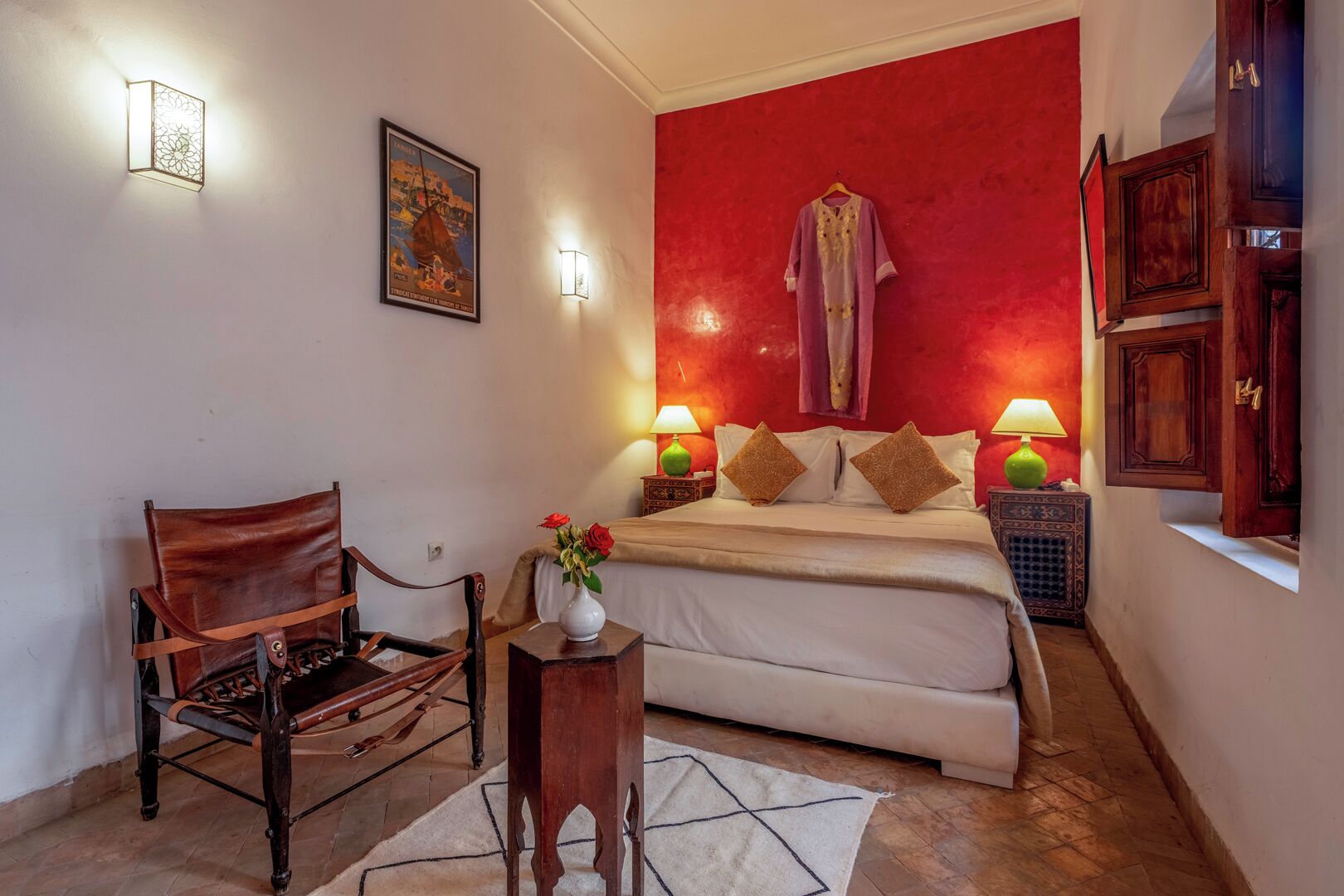 A bedroom with a red accent wall, a bed with two pillows, a chair, a small table with a vase of flowers, and two wall-mounted lamps.