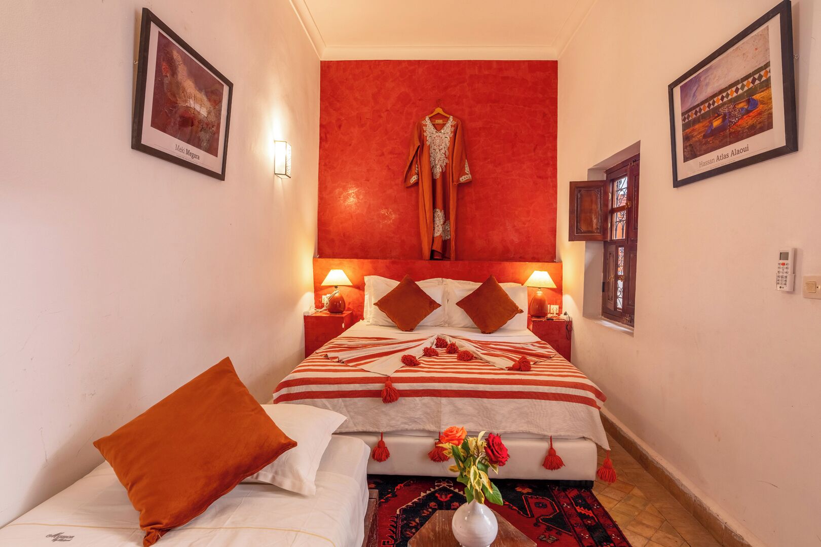 Een kleine hotelkamer met een rode accentmuur, een bed met gestreept beddengoed en decoratieve kussens, een vaas met bloemen en kunstwerken aan de muren.