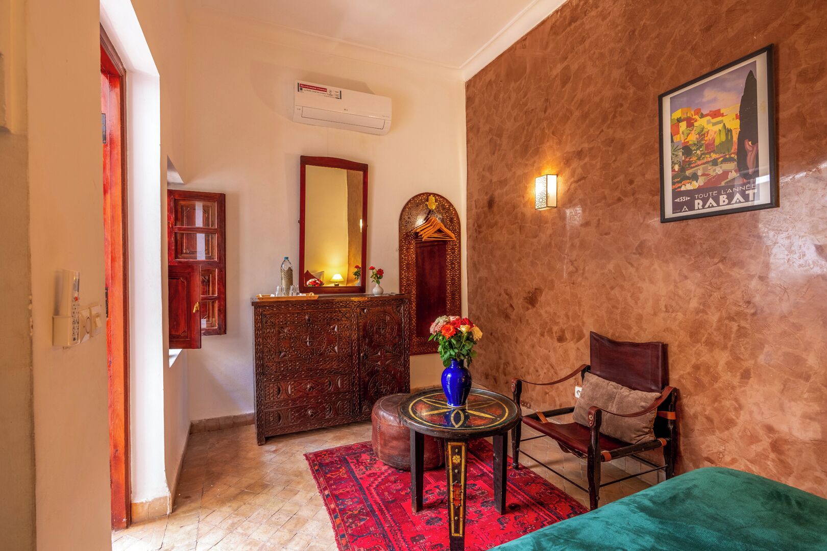 A room with a wooden cabinet, a mirror, a vase with flowers, a bed, and a wall-mounted air conditioner.