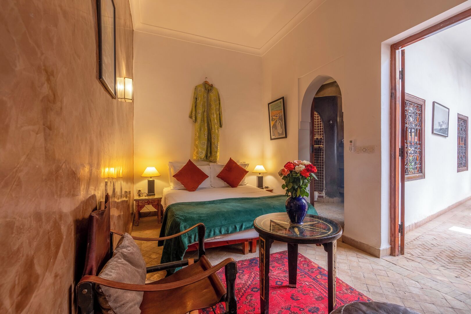Una habitación de hotel con una cama, una silla, una mesita con un jarrón de flores y una alfombra roja.