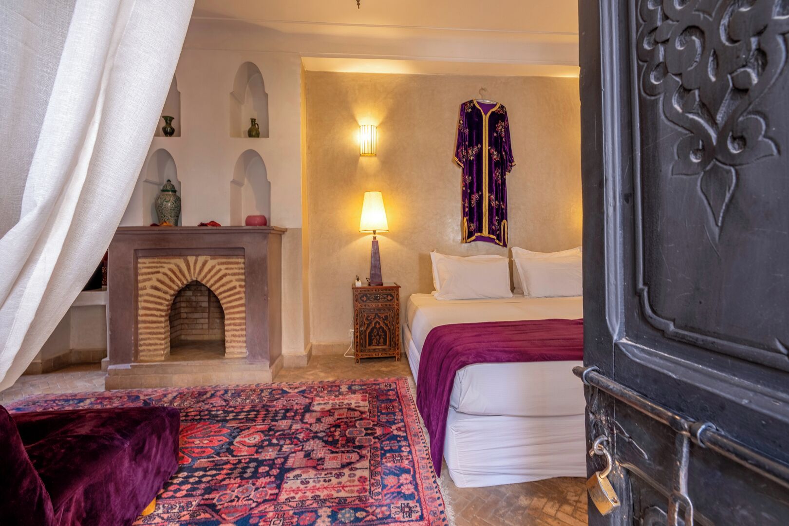 Una habitación tradicional con chimenea, una cama con manta morada, una alfombra estampada y una puerta decorativa.