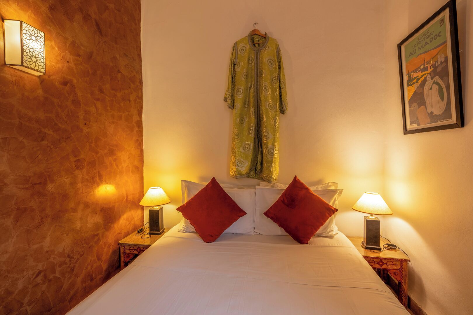 A hotel room with a bed, two bedside lamps, a hanging robe, and a framed picture on the wall.