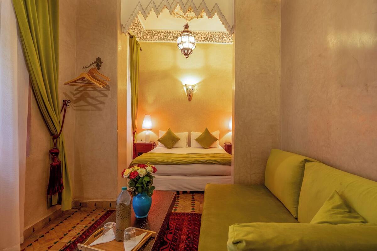 A hotel room with a bed, green curtains, a hanging garment, a vase with flowers, a table with glasses and a tray, and a green sofa.