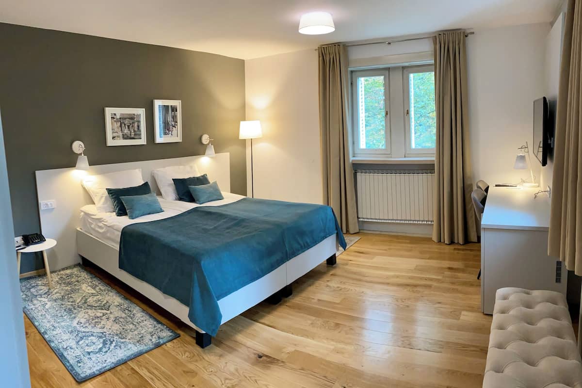 A bedroom with a bed, bedside lamps, a desk with a lamp, a radiator, and a window with curtains.