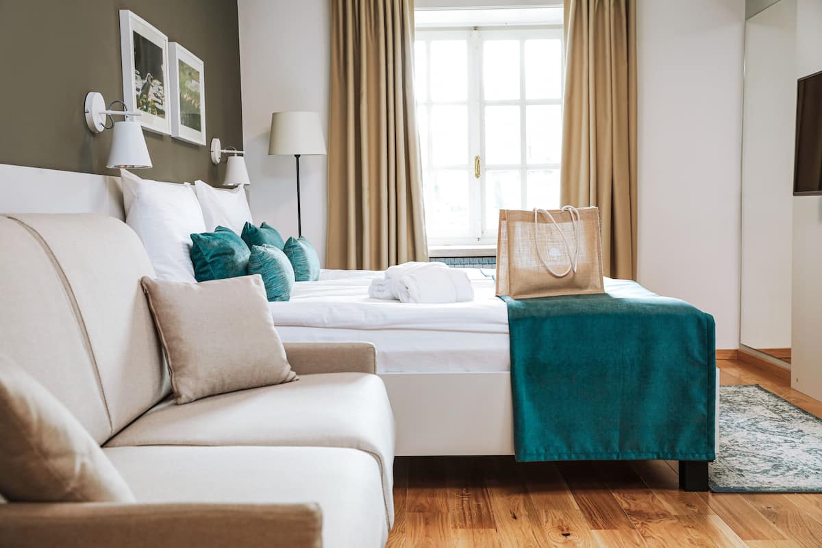 A hotel room with a bed, a sofa, a window with curtains, a lamp, and a bag on the bed.