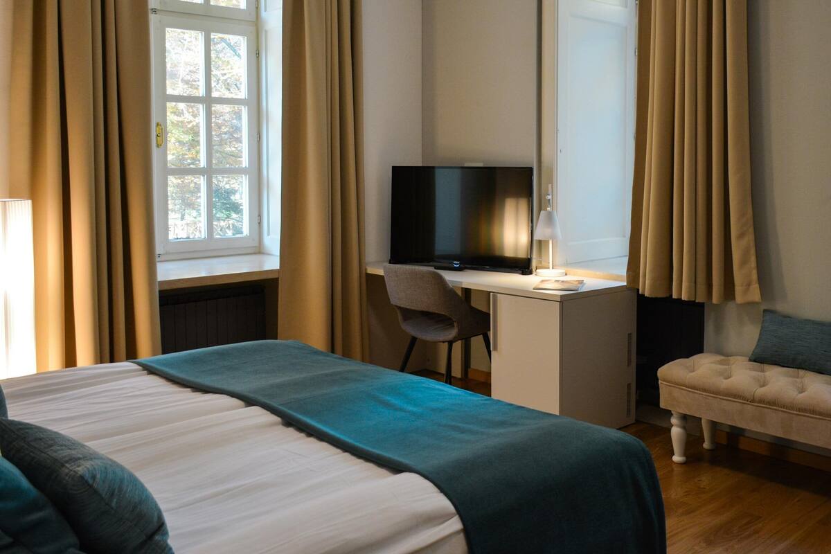 A hotel room with a bed, a desk with a TV, a chair, a sofa, and a window with curtains.