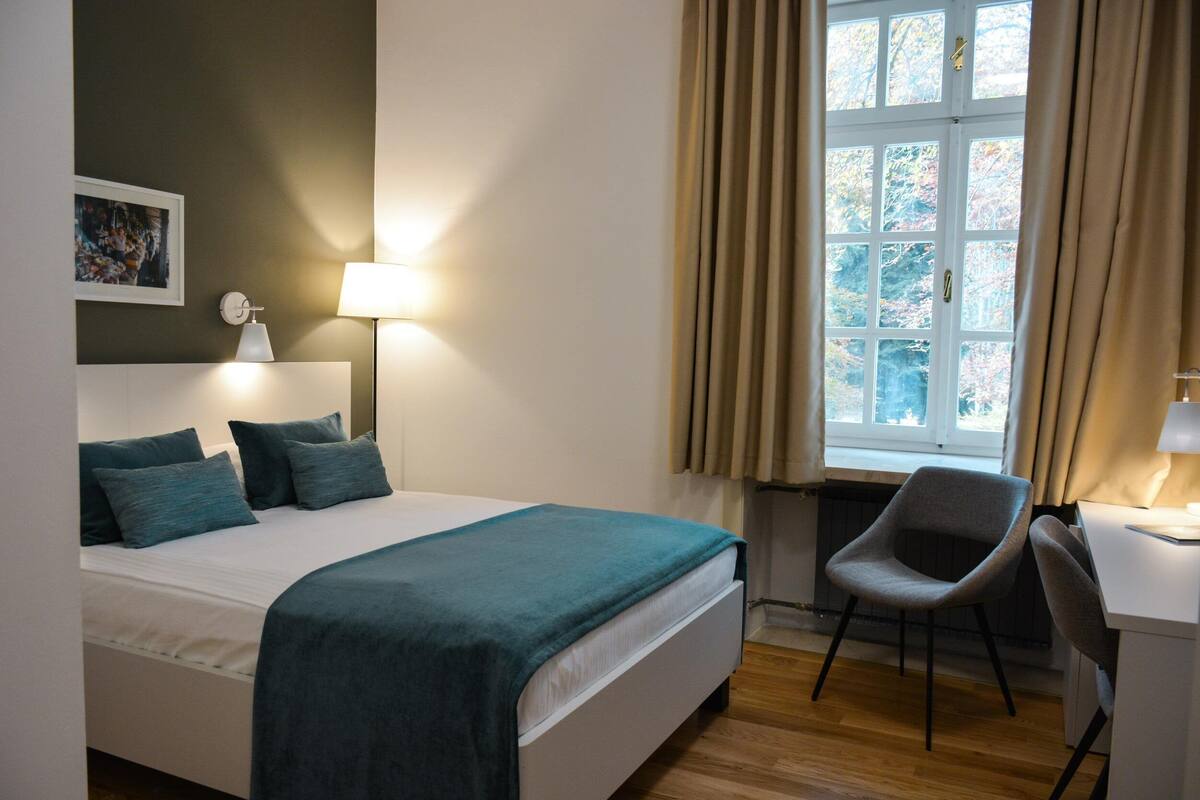 A bedroom with a bed, a desk, a chair, a lamp, and a window with curtains.