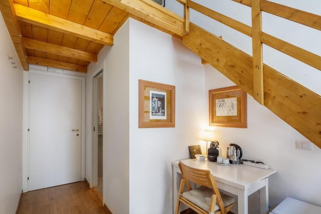 Una habitación con techo de madera, paredes blancas, una puerta blanca, una pequeña mesa con una silla y cuadros en la pared.