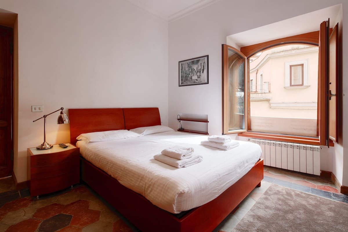 A bedroom with a large bed, a nightstand, a lamp, a radiator, and a window with an open door.
