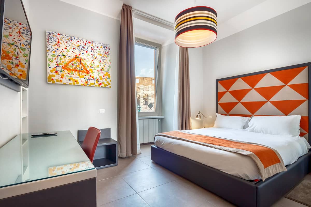 A modern hotel room with a glass desk, a bed with an orange geometric headboard, and a large abstract painting on the wall.