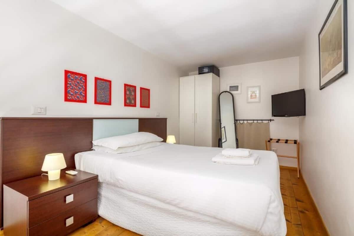 A hotel room with a large bed, a wardrobe, a mirror, a TV, and artwork on the wall.
