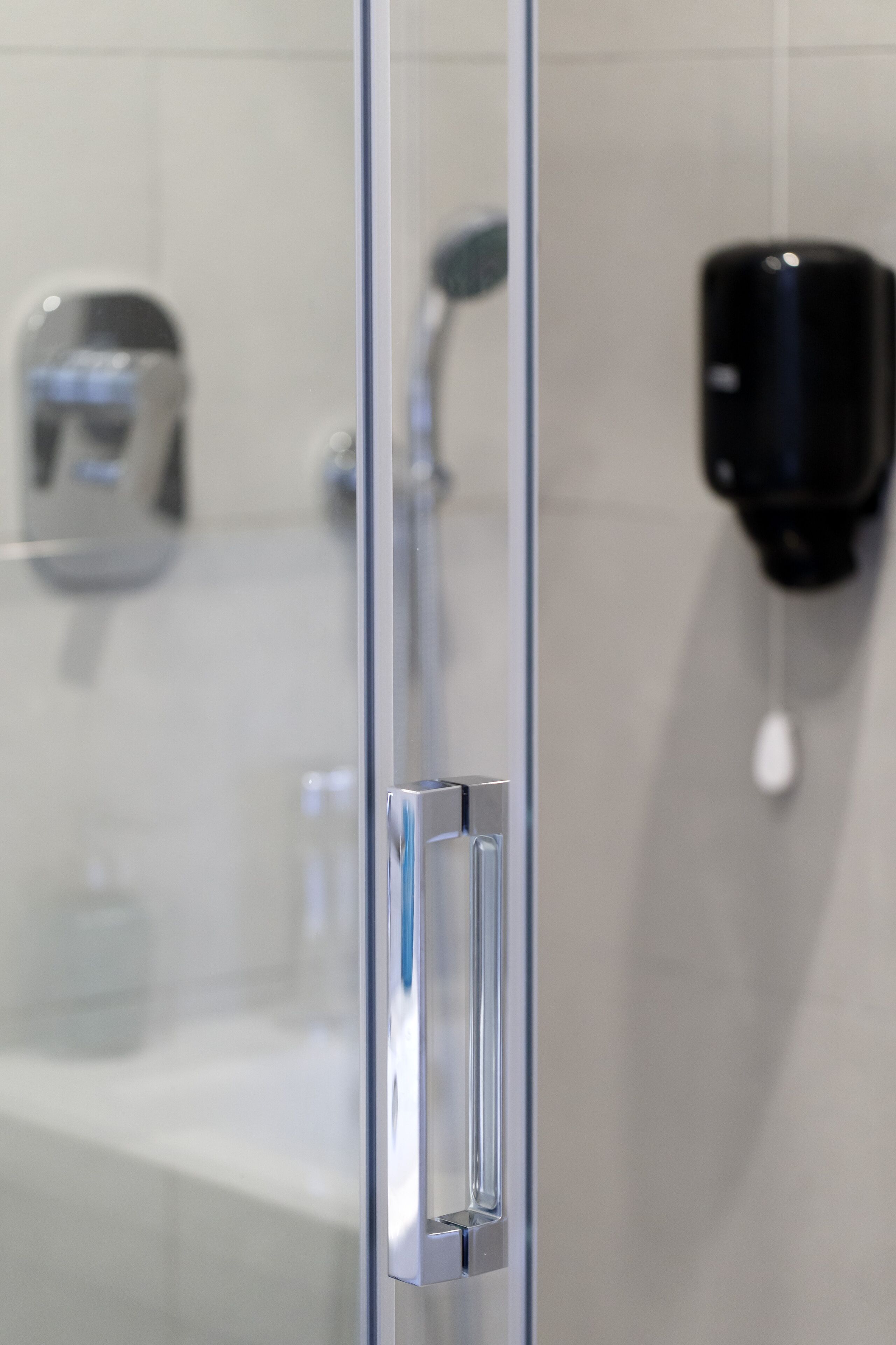 A glass shower door with a chrome handle, a wall-mounted soap dispenser, and a visible showerhead.