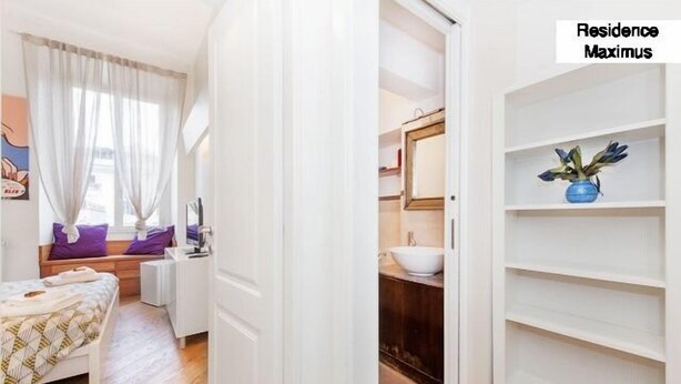 A bedroom with a bed, a window with curtains, a bedside table, and a wall-mounted TV. - Maximus (Rome)