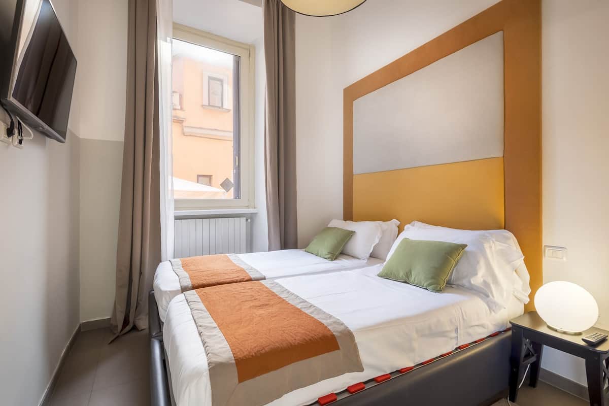 A hotel room with two beds, a TV mounted on the wall, a bedside table with a lamp, and a window with curtains.