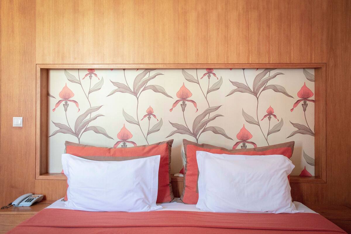 A neatly made bed with white and red pillows, a wooden headboard with floral wallpaper, and a bedside phone on the floor.