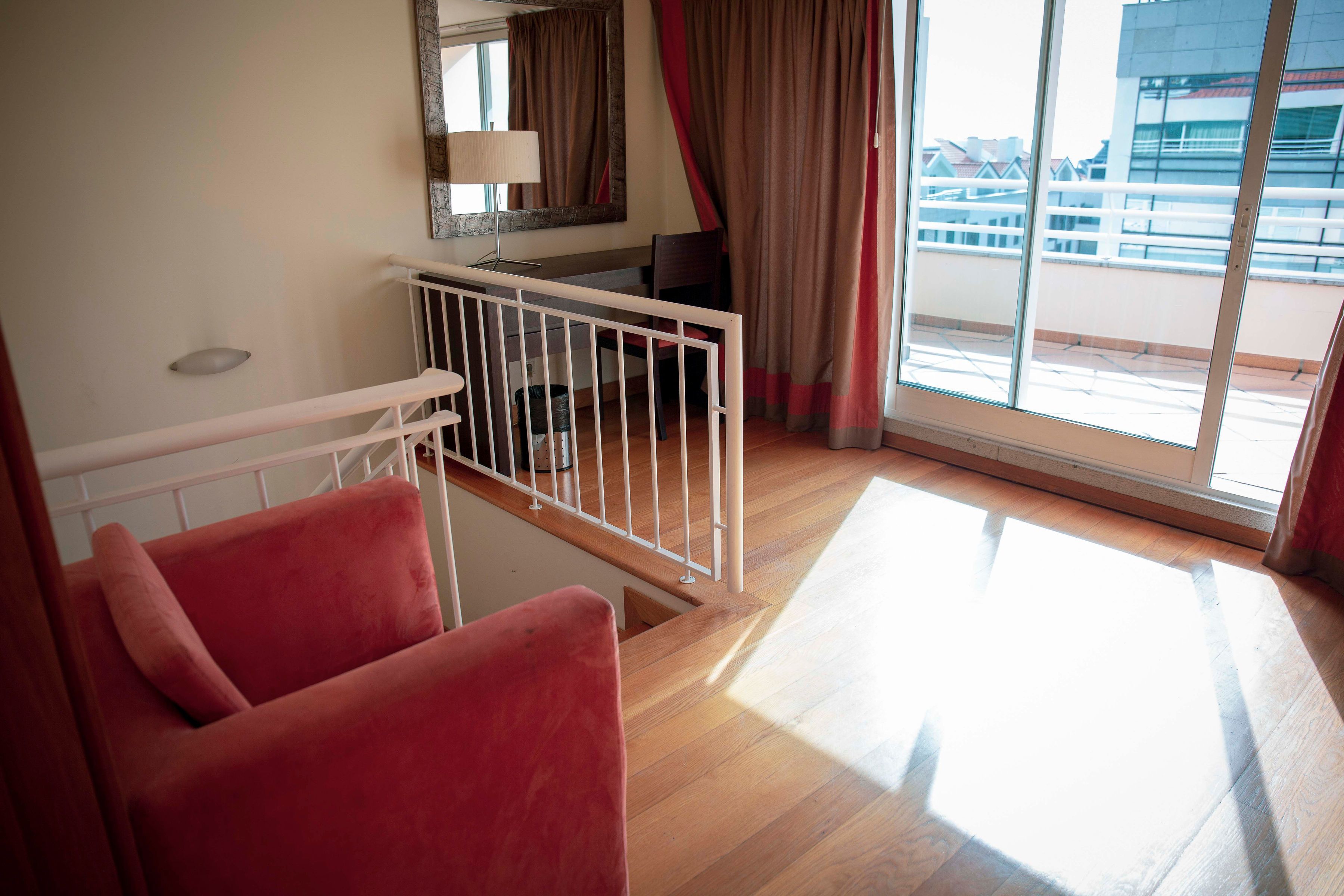 Habitación de hotel con suelo de madera, una silla roja, barandal blanco, escritorio, lámpara y un ventanal con cortinas.