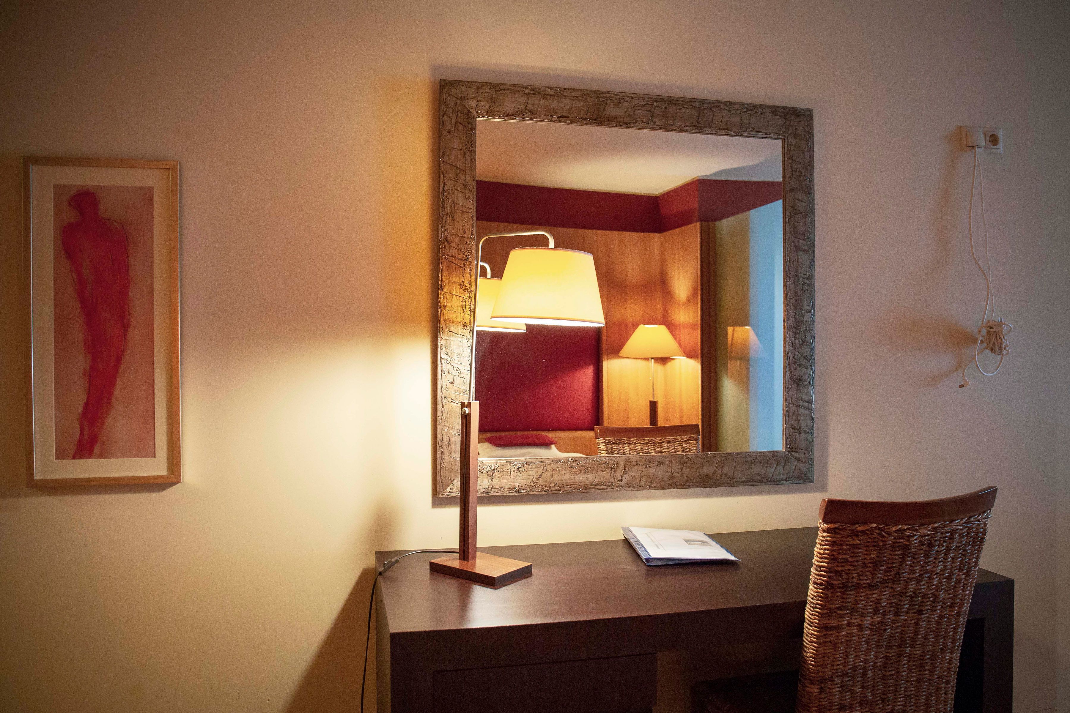 A hotel room with a desk, a lamp, a mirror, and a framed artwork on the wall.