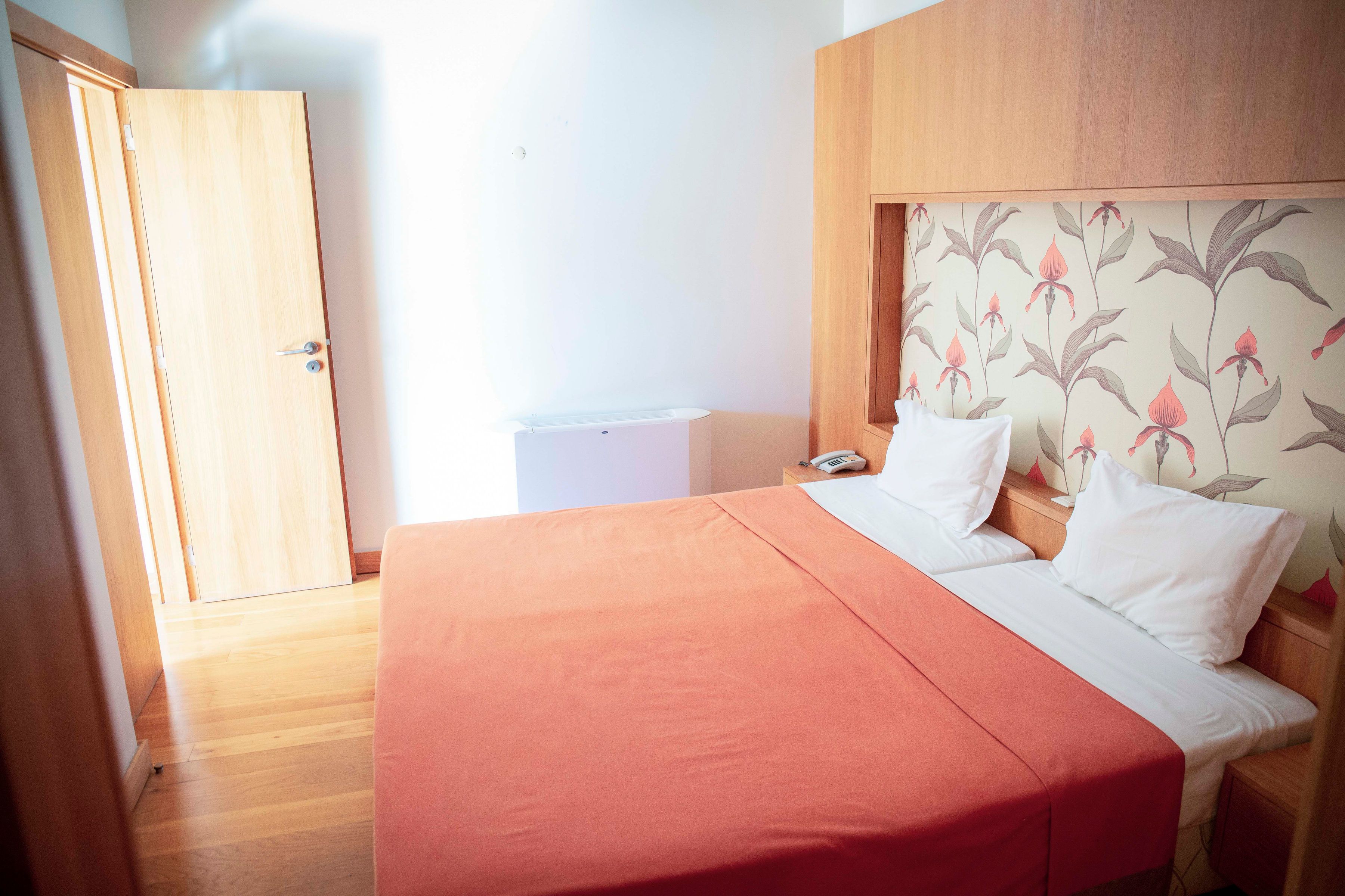 A bedroom with a wooden bed frame, a red bedspread, white pillows, a nightstand with a phone, and a wall with a floral wallpaper pattern.
