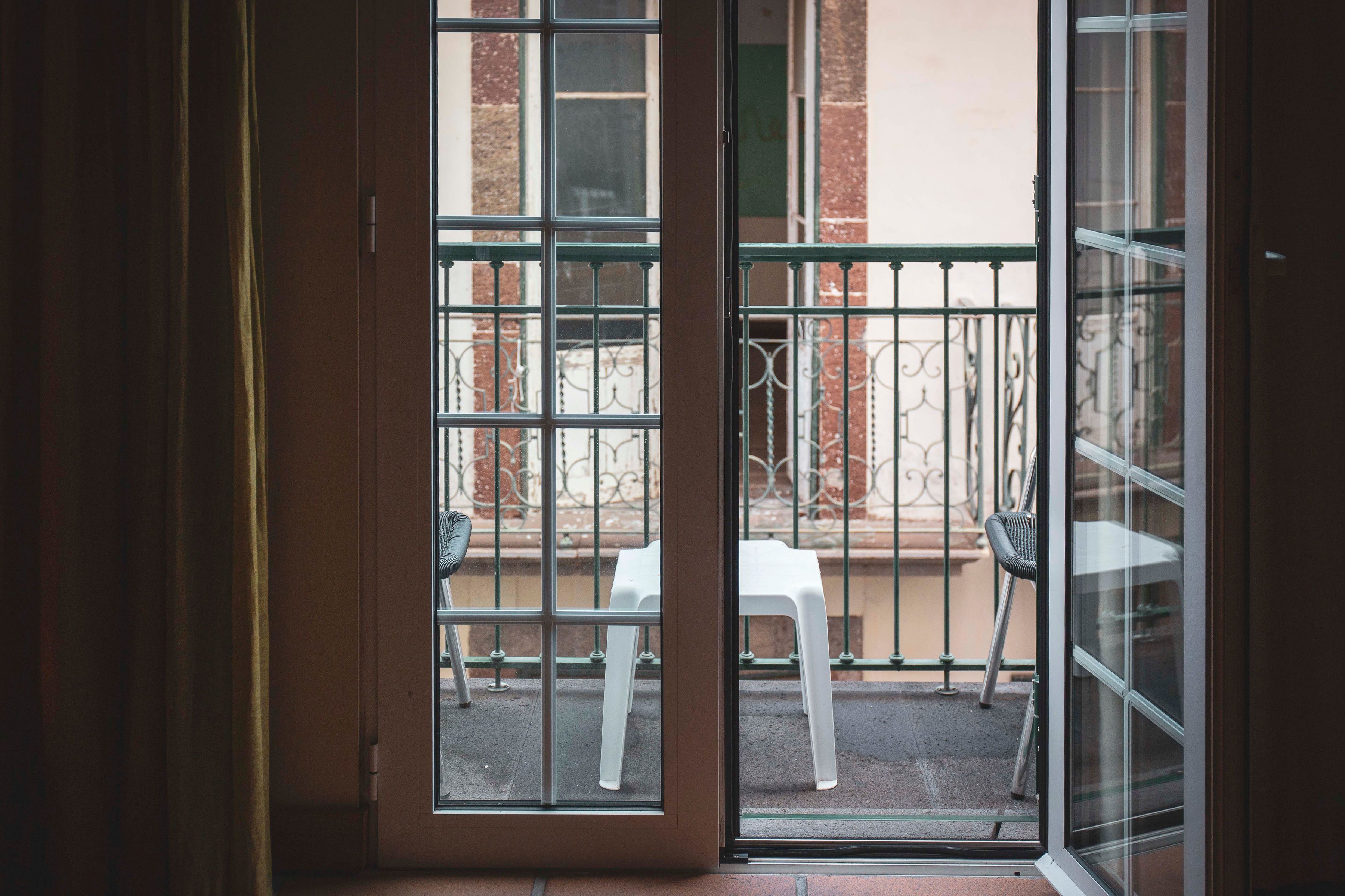 Blick durch eine Glastür auf einen Balkon im Freien mit einem weißen Stuhl und einem Metallgeländer.