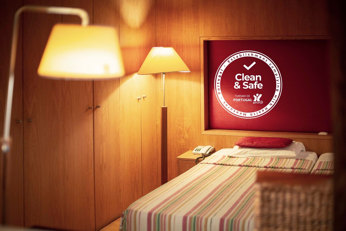 A hotel room with a bed, a nightstand, a lamp, and a wall-mounted sign reading "Clean & Safe" with the logo of Turismo de Portugal.