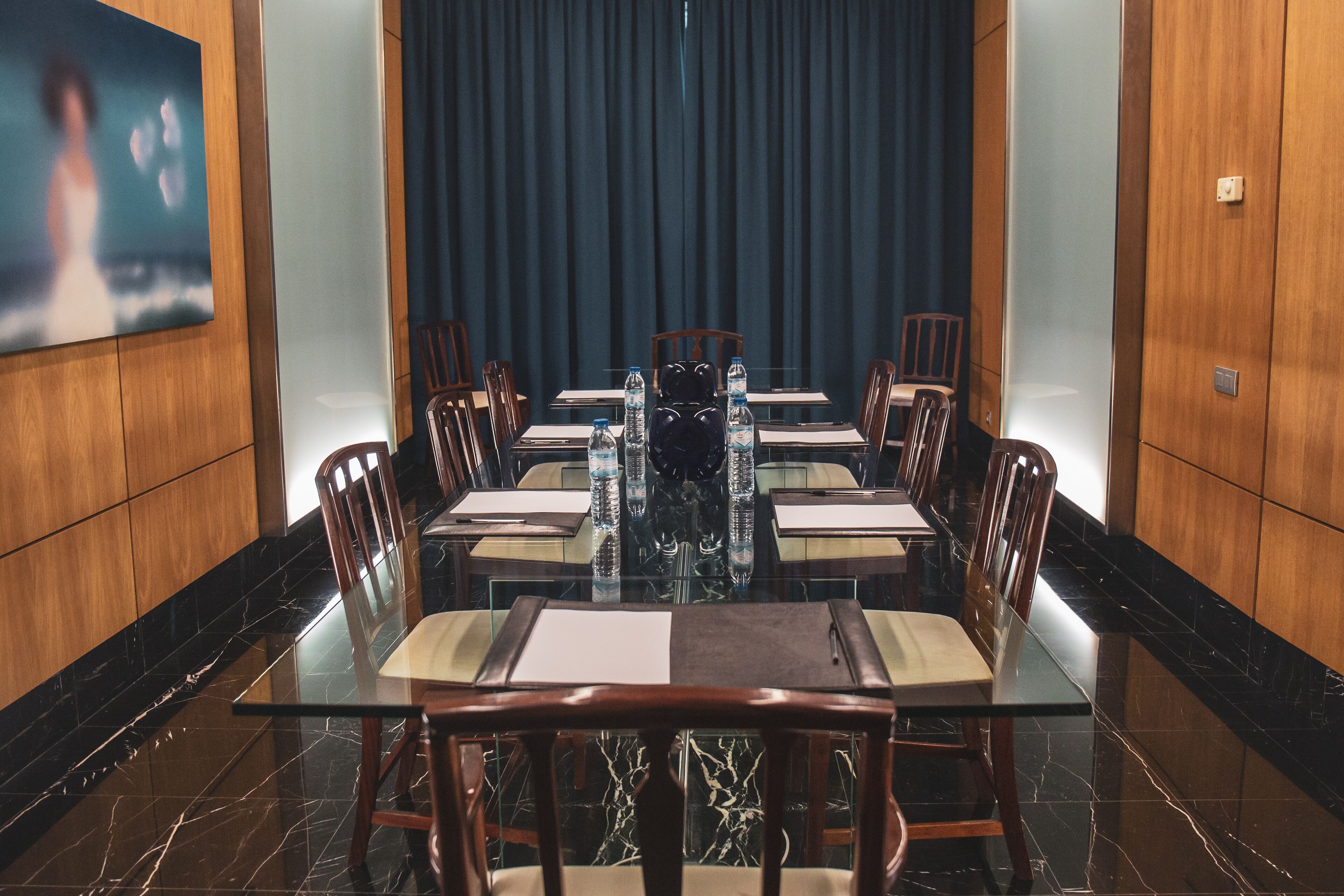 Een vergaderruimte met een glazen tafel, houten stoelen en een groot schilderij aan de muur.