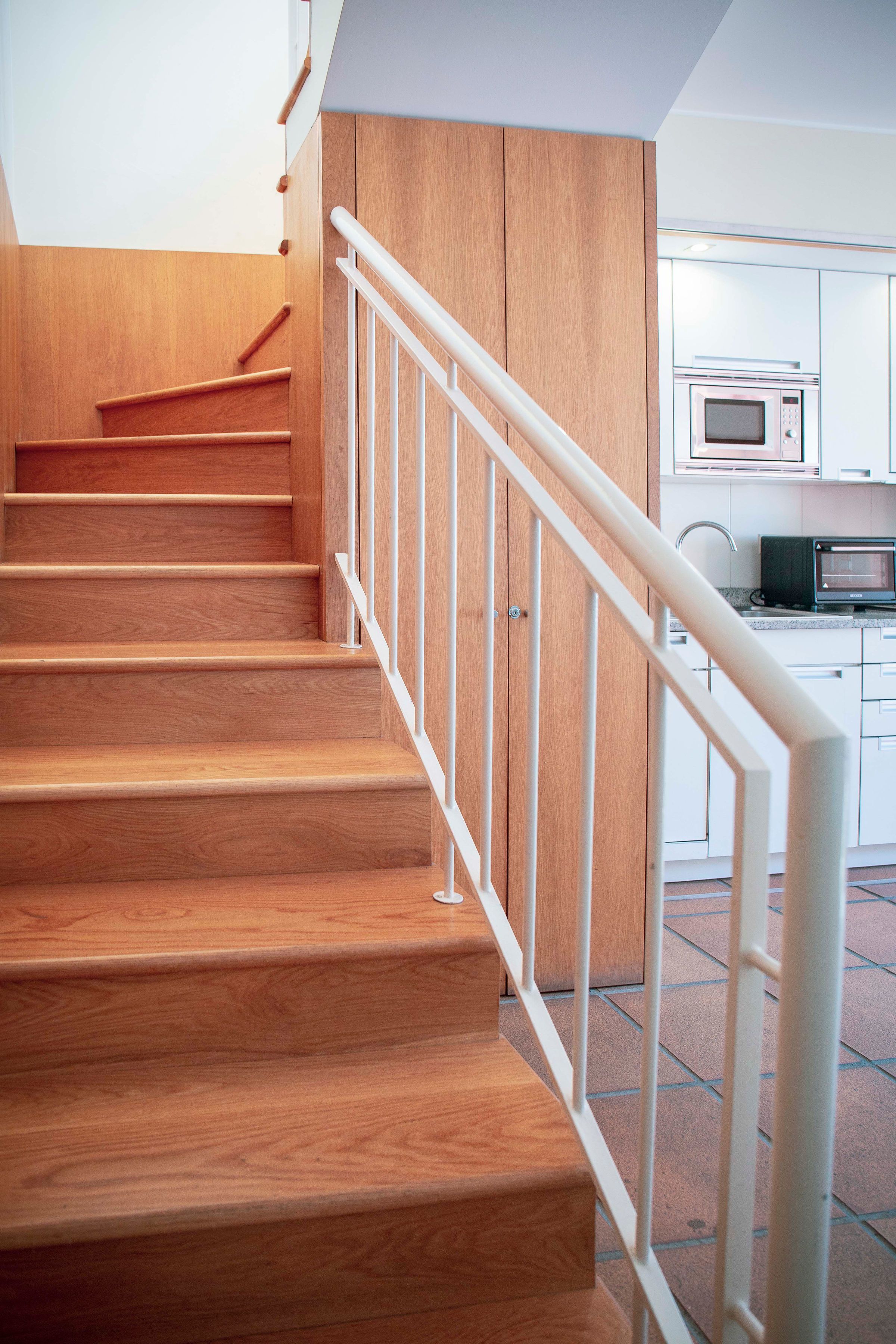 A wooden staircase with white railings leading to a kitchen area with modern appliances.