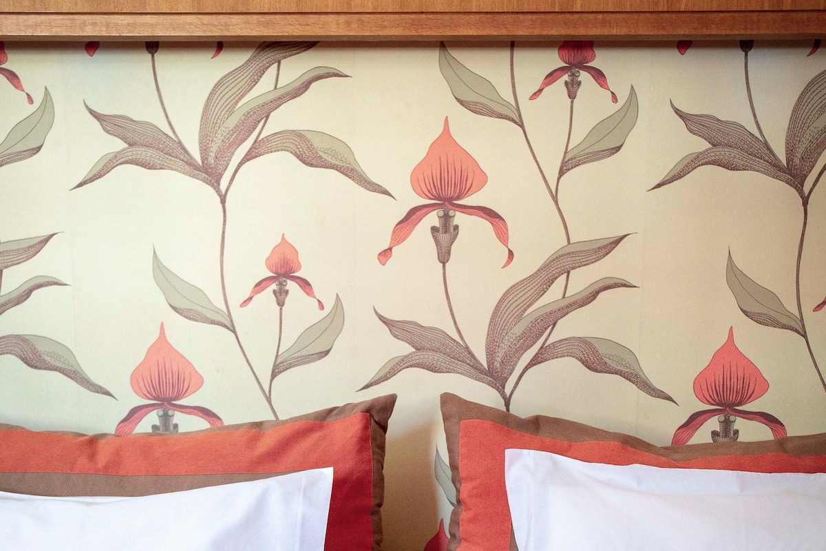 A bed with red bedding and white pillows, a wooden headboard, and a floral wallpaper pattern.