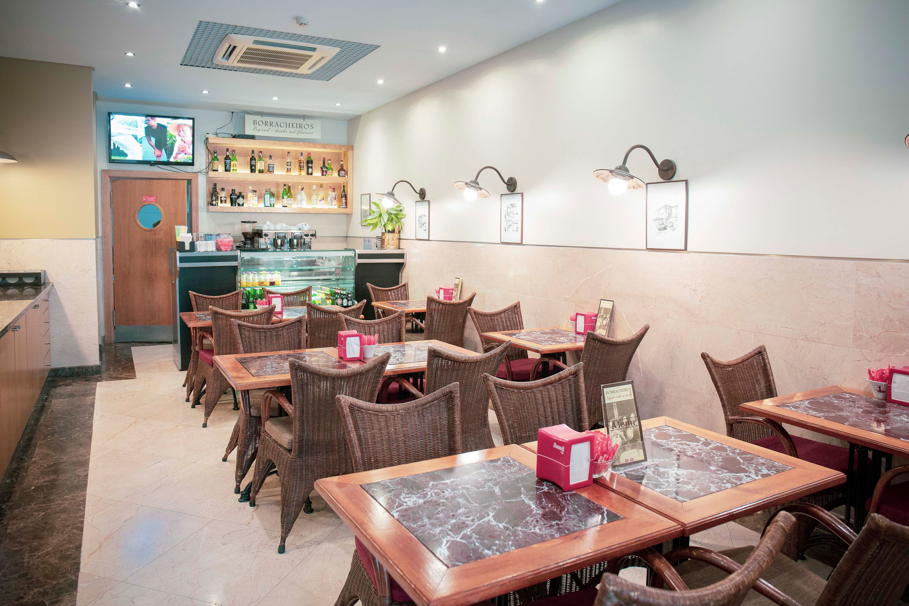 A restaurant interior with wicker chairs, marble-topped tables, a bar area with shelves of bottles, and a television screen.